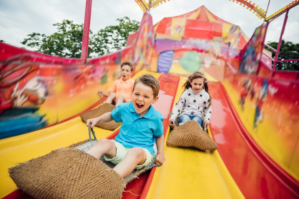 Children having fun sliding down a yellow and red slide while sitting in burlap sacks at a fair.