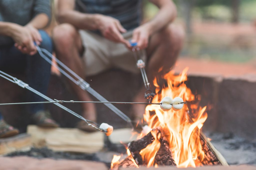 Close-up shot of a campfire with four metal skewers roasting marshmallows on a summer evening.