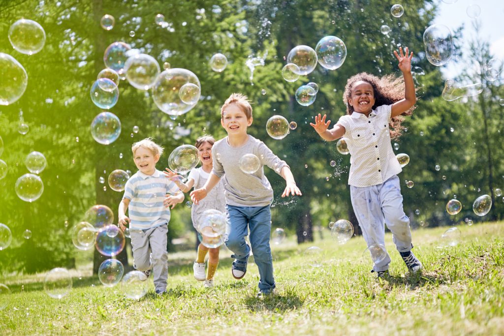 Group of little friends with toothy smiles on their faces enjoying warm sunny day while participating in soap bubbles summer birthday party.