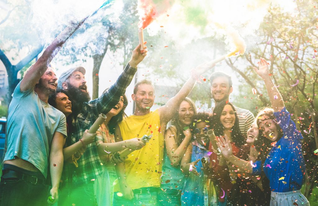 Happy friends having fun at summer birthday party with multicolored smoke bombs outdoors.