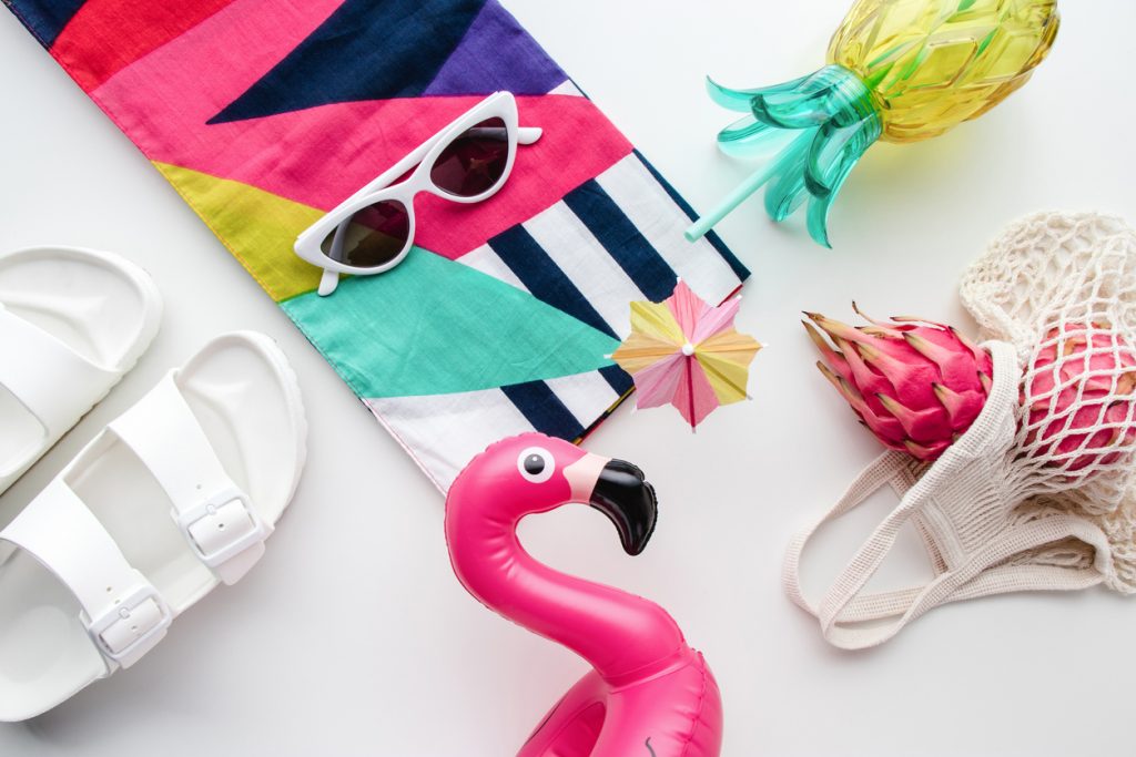 Flat lay of summer birthday party vibes concept with colorful travel fashion items, sunglasses, scarf, pink dragon fruit, flamingo inflatable drink holder, pineapple straw tumbler bottle, net bag and sandal on a white background.