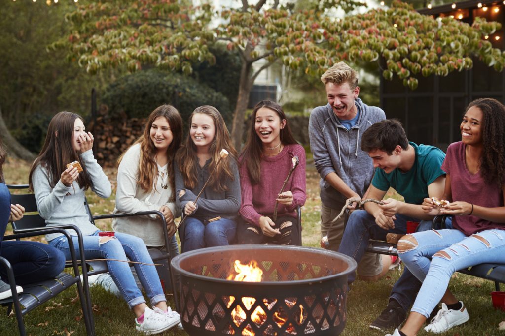 Teenage friends sit round a fire pit toasting marshmallows.
