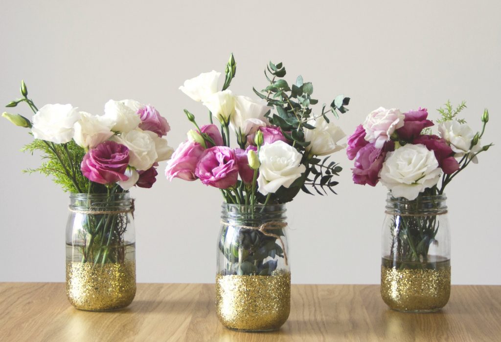 Bouquets of flower arrangements in gold glass jars as centerpieces on a wooden shelf or table.
