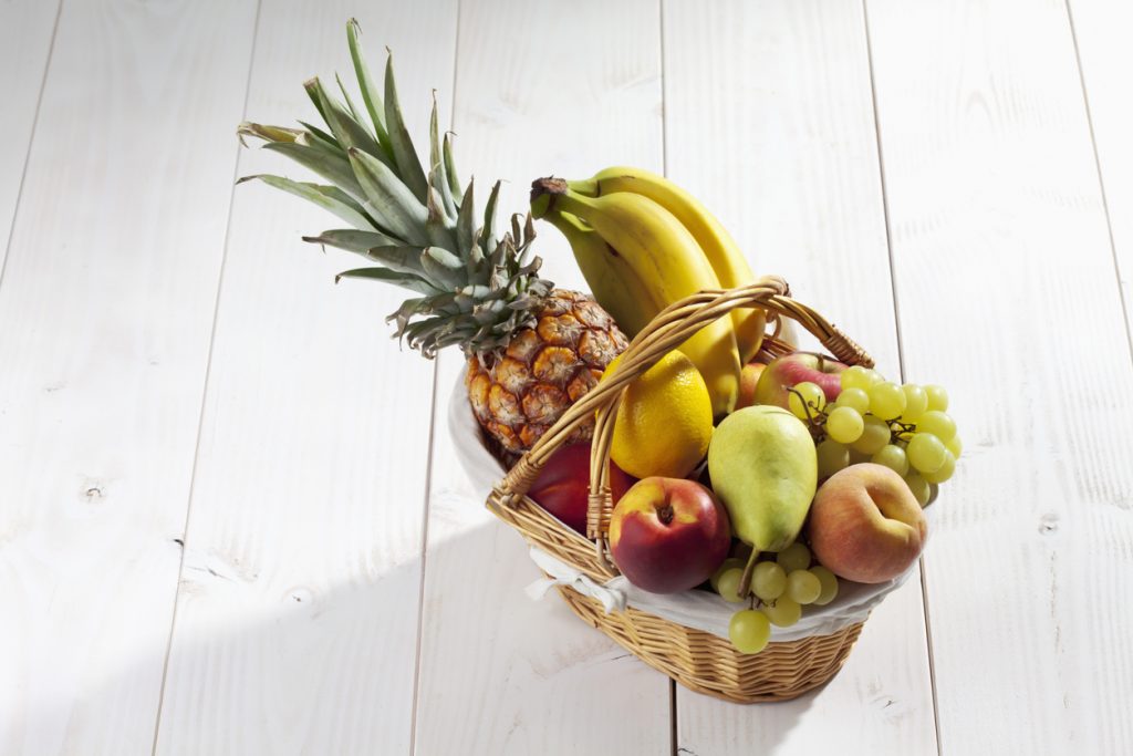 Fruit basket with pineapple, bananas, lemon, apple, peaches, grapes on white wooden background.