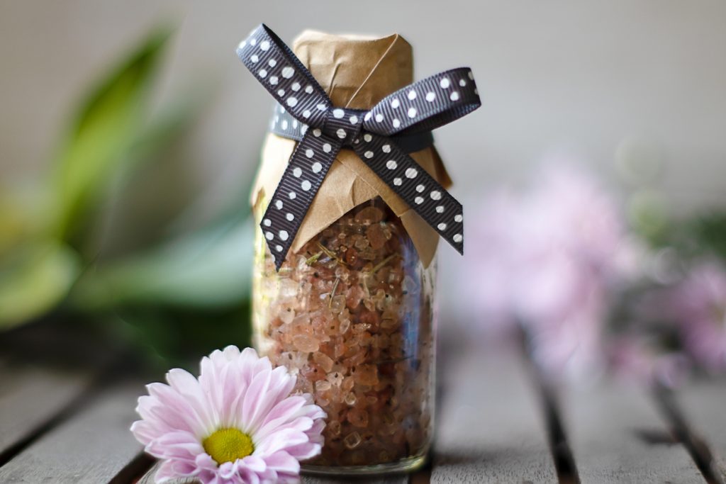 Pink organic sea salt in glass bottle decorated with pink pastel daisy flower on wooden table.