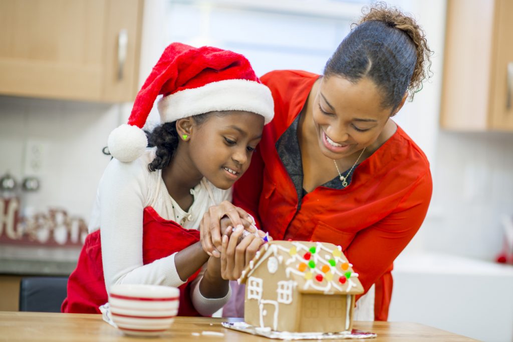 A mother and daughter are decorating a gingerbread house at home in their kitchen.