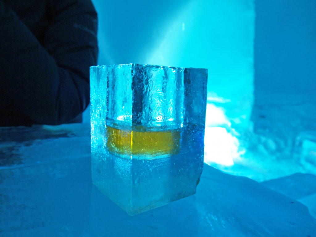 Frozen Shot glass with whiskey made of ice, shaped form frozen water in dark blue ice bar environment.