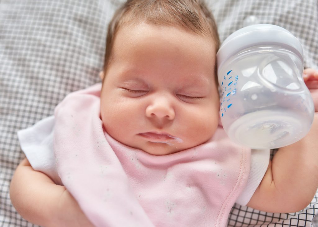 Cute baby girl sleeping, milk bottle near her face with a pink bib.
