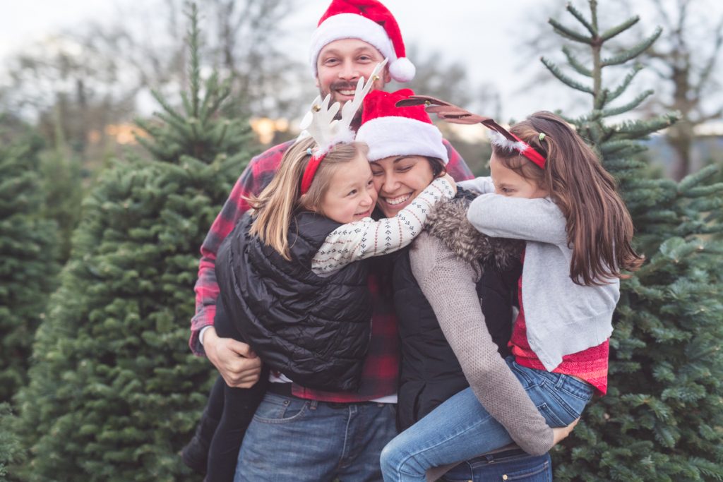 A stylish Santa hat wearing couple pose for a photo with their girls at the Christmas tree farm. Mom is holding one, dad is holding the younger, and they all have big smiles.
