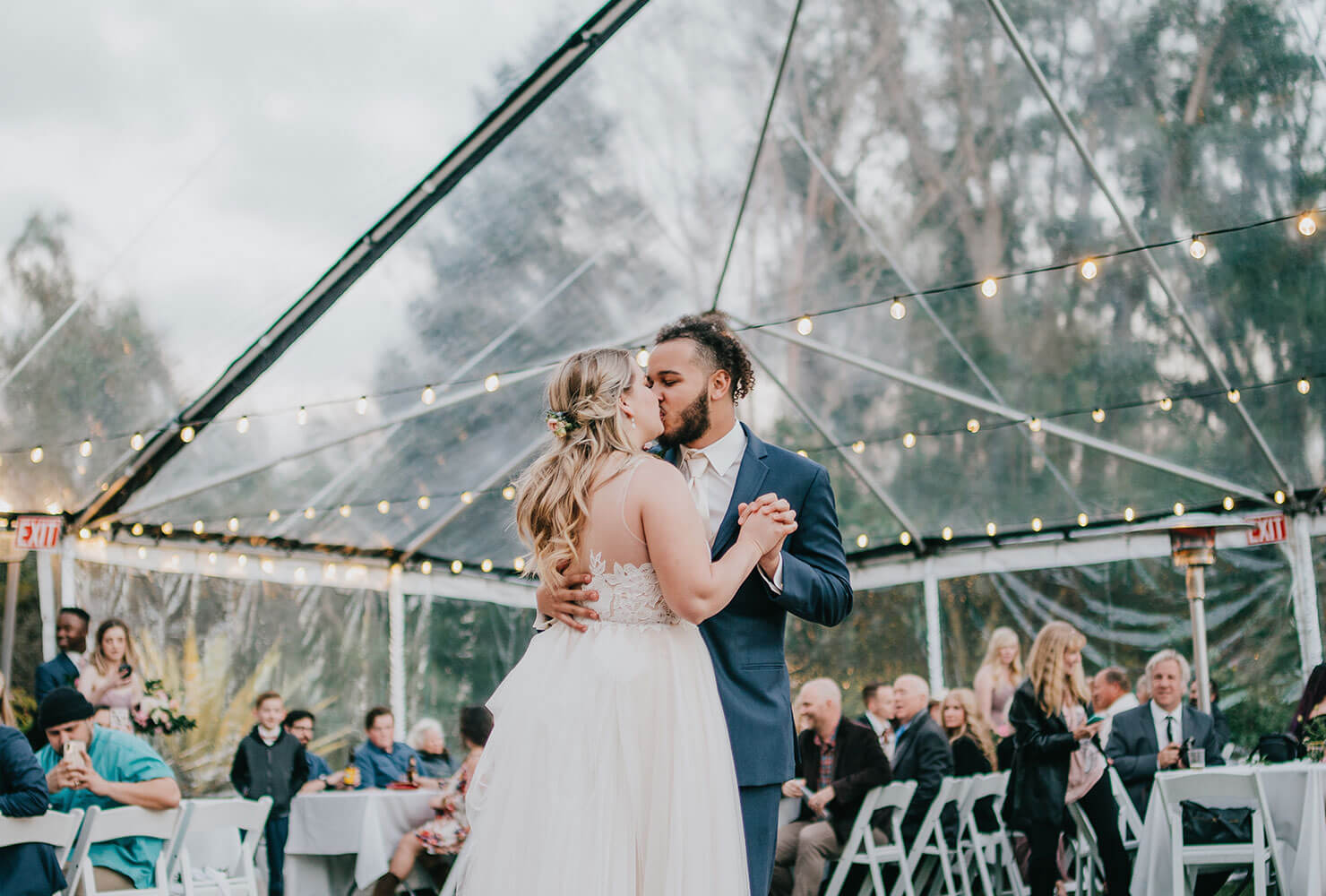 Couple during their first dance under a clear tent surrounded by guests.