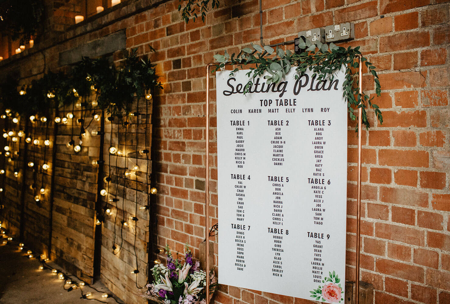Wedding seating chart on a brick wall decorated with vines and market lights.