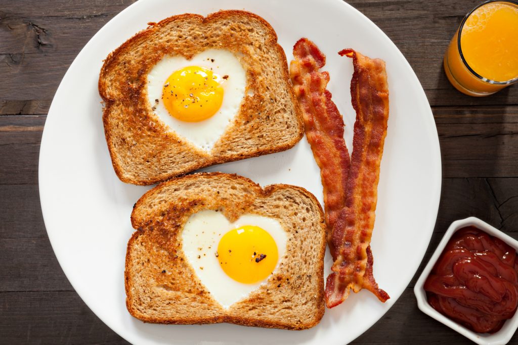 An overhead close up shot of heart shaped eggs inside toast, a couple of bacon strips and fresh squeezed orange juice