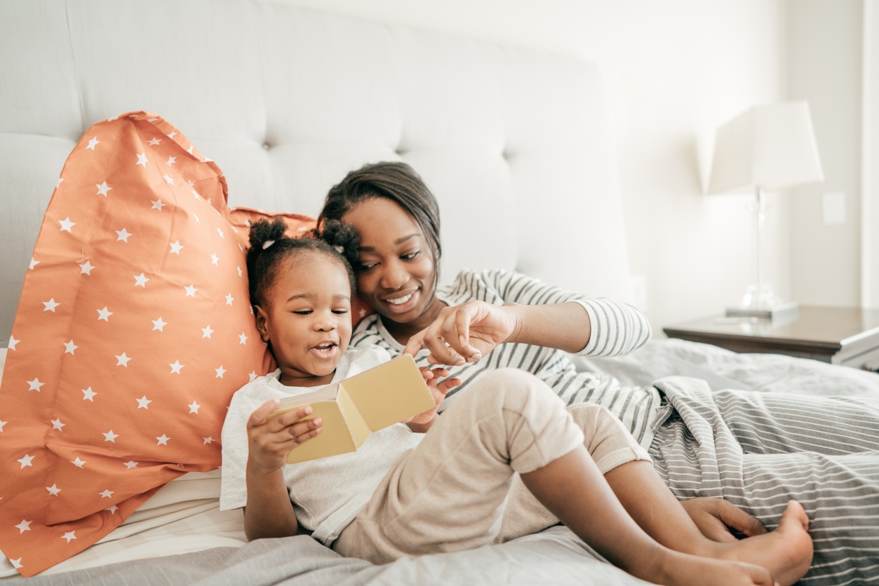 Mom and her toddler daughter reading a small book in bed.